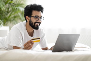 Çevrimiçi ödemeler. Evde yatakta dinlenirken dizüstü bilgisayar ve kredi kartı kullanan gülümseyen Hintli adam, rahat yatak odasında dinlenirken bilgisayarla internet alışverişi yapan mutlu genç doğu erkeği,