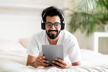 Yatakta dijital tabletle rahatlayan neşeli Hintli genç adam, kablosuz kulaklıklar takan, rahat yatak odasında dinlenen, internete göz gezdiren ya da internetten alışveriş yapan genç adam.
