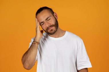 Bin yıllık mutsuz, beyaz tişörtlü beyaz bir adam. El ele tutuşmuş, acıdan muzdarip, turuncu stüdyo arka planında izole edilmiş. Hata, stres, baskı ve sağlık sorunları, migren
