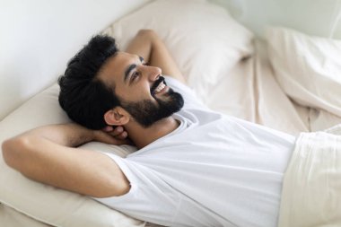 İyi sabahlar. Mutlu Hintli genç bir adamın portresi, rahat bir yatakta uyanır, yakışıklı, gülümseyen, elleri başının arkasında uzanan, yatak odasında dinlenen, boş zamanlarının tadını çıkaran, yakın çekim yapan...