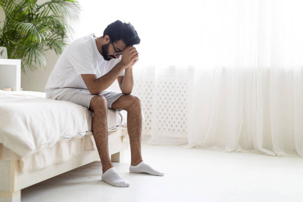 Портрет подавленного молодого индийца, сидящего дома на кровати, расстроенного восточного парня тысячелетия, зарывающего голову в руках, страдающего жизненными проблемами или психическим срывом, чувствующего отчаяние, копирующего пространство