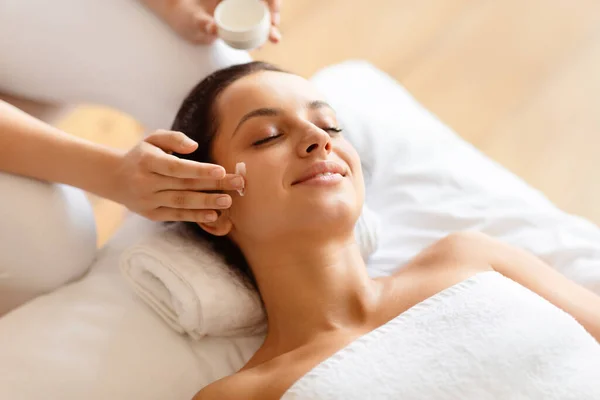 Facial Spa. Young Woman Enjoying Relaxing Face Massage While Masseur Applying Moisturizer Cream On Face Skin, Lying Wrapped In Towel In Luxury Spa, Posing With Eyes Closed. Skincare Concept