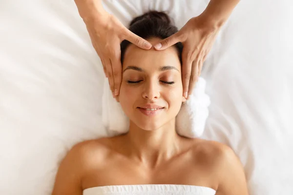 Spa And Relax. Young Lady Receiving Head Massage Enjoying Beauty Ritual Lying With Eyes Closed On Bed Indoor, Top View. Masseur Massaging Ladys Forehead And Face For Relaxation