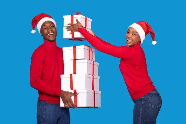 Christmas Presents. Happy Young Black Woman Putting Another Xmas Gift To Stack In Man Hands, Couple Posing With Many Wrapped Gifts Wearing Santa Hats On Blue Background. Winter Holidays Greetings