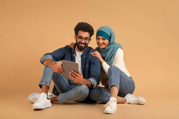 Happy Young Arab Couple With Digital Tablet Relaxing On Floor Over Beige Studio Background, Smiling Muslim Man And Woman In Hijab Using Modern Gadget For Online Shopping Or Browsing Internet