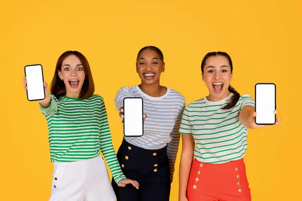 Shopping mobile app concept. Cute pretty stylish young multiethnic women showing phones with white empty screen, isolated on yellow background, purchasing online, sale season, e-commerce