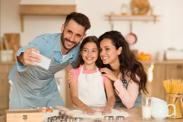 Happy family baking cookies and making selfie on cellphone parents embracing daughter, standing in modern kitchen at home on weekend. Pastry recipes concept