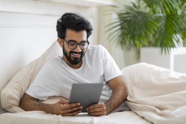 Yakışıklı Hintli adam yatakta dinlenirken dijital tablet üzerinde interneti tarıyor, evde modern Gadget kullanan gülümseyen genç adam, online alışveriş yapıyor ya da yakın çekim filmleri izliyor,