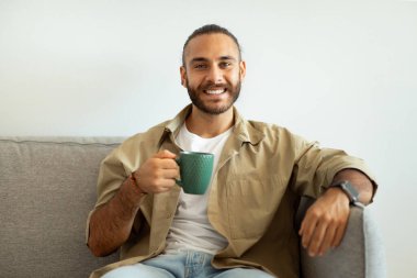 Oturma odasındaki koltukta oturan, elinde kahve fincanı tutan mutlu, yakışıklı, sakallı genç adam, kameraya gülümsüyor, ev içi, fotokopi odası. Pozitif adam sıcak aromatik çaydan hoşlanıyor.