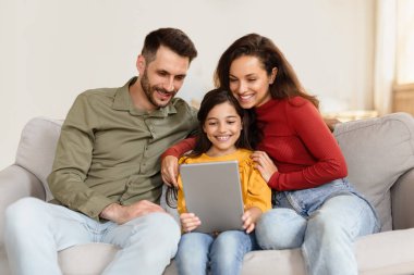 Aile zamanı. Mutlu İspanyol anne, baba ve kız dijital tablet kullanıyor, evdeki kanepede internette geziniyor, kapalı mekandaki bilgisayarla birlikte oturuyor. Gadget ve modern online eğlence konsepti