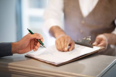 Erkek el imzalarken otel resepsiyonunda kayıt yaptıran üniformalı resepsiyonist, imzalanacak yeri gösteren, parmak işareti yapan kapalı mekan. Kırpılmış Vuruş, Seçici Odaklanma