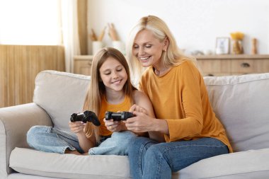 Neşeli, güzel, sarışın, beyaz, okul çağındaki kız büyükannesine bilgisayar oyunu oynamayı gösteriyor. Evde kanepede oturan mutlu büyükanne ve torun, ellerinde joystickler konsolu