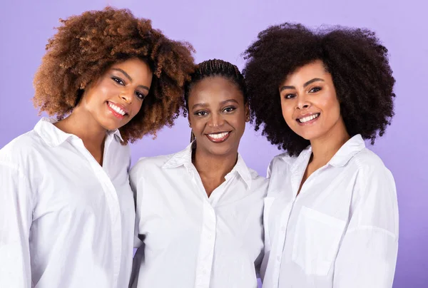 Female Friendship. Studio Portrait Of Three African American Ladies In White Shirts, Hugging Showing Their Unity, Expressing Good Vibes Smiling At Camera Together Over Purple Background