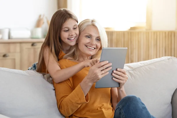 Positive joyful attractive blonde elderly woman 60s having fun with her grandchild pretty teen aged girl at home, sit on couch in living room, using digital tablet, play mobile game, using nice app