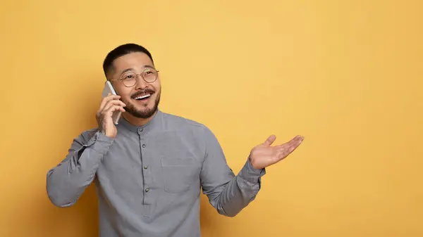 モバイルコミュニケーション エキサイティングな若いアジア人男性は 携帯電話で話し 離れて見て 笑顔ミレニアルガイイイイイイイイイイ黄色い背景の上に立っている間 楽しい会話を楽しんでいます コピースペース — ストック写真