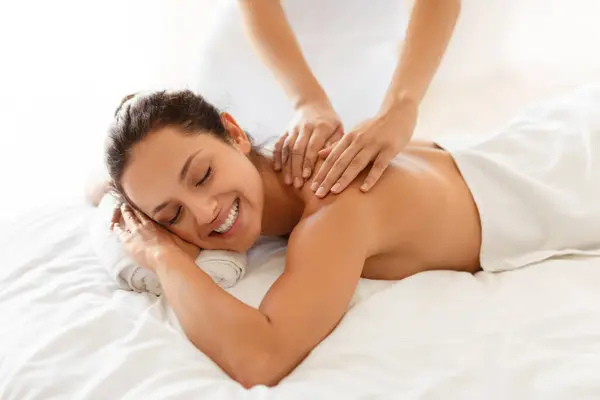 Body Care And Spa. Young European Lady Enjoying Massage, Relaxing With Eyes Closed, Lying On Bed Indoors, While Masseuse Massaging Her Shoulders For Relaxation And Wellness. Cropped
