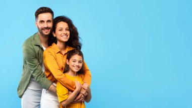 Sevgi dolu Avrupalı aile genç karı koca ve küçük kızı ile birlikte mavi stüdyo arka planında kameraya sarılıyor ve gülümsüyor.