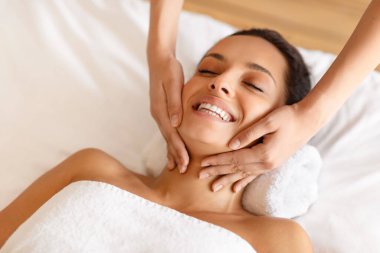 Gülümseyen, rahatlatan, yatakta uzanan, masaj yapan, masaj yapan, kaplıcada yüzüne ve boynuna masaj yapan, gözleri kapalı poz veren kadın. Rahatlama, Güzellik ve Sağlık Konsepti