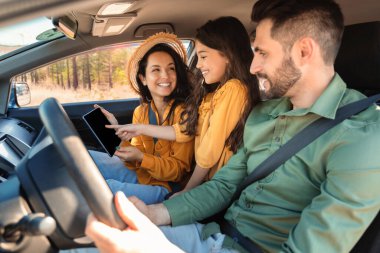 Mutlu bir aile, kızıyla birlikte arabada oturuyor ve dijital tablet kullanıyor, online haritaları kontrol ediyor, ebeveynler ve kadın çocuk bir sonraki seyahat planını birlikte yapıyor.