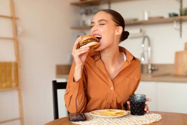 Abur cubur. Lezzetli hamburger yiyen, lezzetli, sağlıksız fast food tutan ve yiyen bir kadın. Mutfaktaki masada oturuyor, boş yer, pankart.