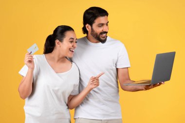 Bin yıllık gülümseyen Arap kocası dizüstü bilgisayarı ve kredi kartı ile polisi işaret eden karısı sarı stüdyo arka planında izole edilmiş. Aile, internetten alışveriş, reklam ve teklif, satış.