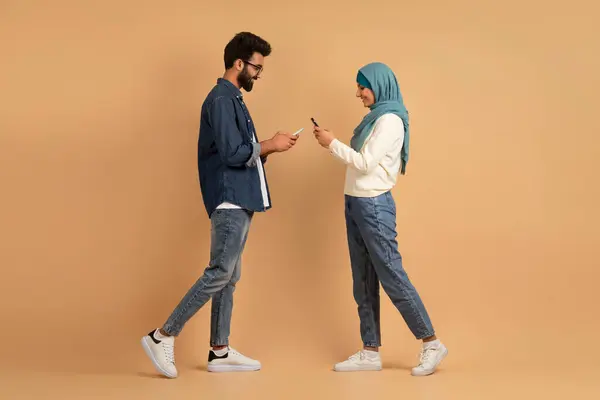 Muslim Dating Apps. Happy Young Islamic Couple With Smartphones In Hands Walking Toward Each Other, Romantic Arab Man And Woman In Hijab Enjoying New Application, Posing On Beige Background