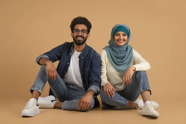 ベージュスタジオの背景に床に座っているヒジャブの若いアラビア人男性と女性の肖像画 一緒にリラックスする幸せなムスリムの肖像画 笑顔と見るカメラ 無料スペース — ストック写真