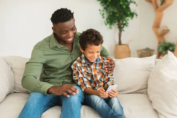 小男孩和他的黑人爸爸一起用手机在网上玩游戏 在家里上网浏览沙发放松 父亲在孩子使用小工具的时候拥抱男孩 — 图库照片