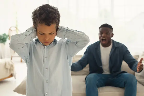子どもに対する家庭内暴力 マッド アフリカ系アメリカ人の父親は 子供が家で耳を覆っている間 怖がった子供の息子に叫びました 父親とのクォーレル中に不幸な小さな男の子に選択的な焦点 — ストック写真
