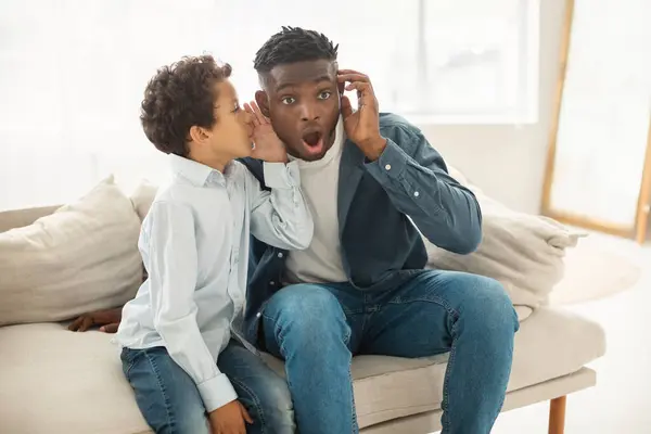 Little son whispering in fathers ear secrets, rumors and news sitting on couch at home interior. Overwhelmed black father listens to his kid sharing surprising information. Family communication