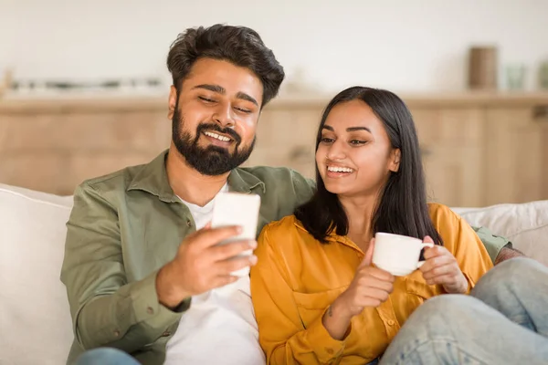 インドのカップルは 携帯電話でソーシャルメディアを共有し ソファーに座っている幸せな配偶者 ガジェットとコーヒーを飲む女性を使用しています スマートフォン画面を見ている女性を受け入れる笑顔ヒンドゥー男性 — ストック写真