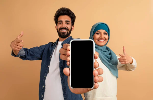 Nice Offer. Smiling Muslim Couple Showing Big Blank Smartphone In Their Hands, Arab Man And Woman In Hijab Demonstrating Copy Space For Mobile Advertisement, Standing On Beige Background, Mockup