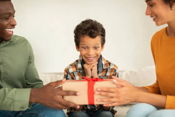 Gift Giving And Celebration. Diverse Parents Surprising Kid Boy With Present, Showing Wrapped Box To Son Celebrating Birthday At Home. Cropped Shot, Selective Focus On Happy Child