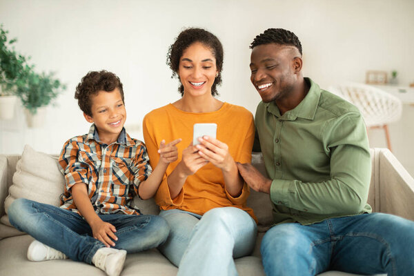 Portrait Of Smiling Multiethnic Family With Little Son Using Smartphone Together At Home, Happy Diverse Parents And Cute Kid Boy Checking New Mobile App While Relaxing On Couch Indoor