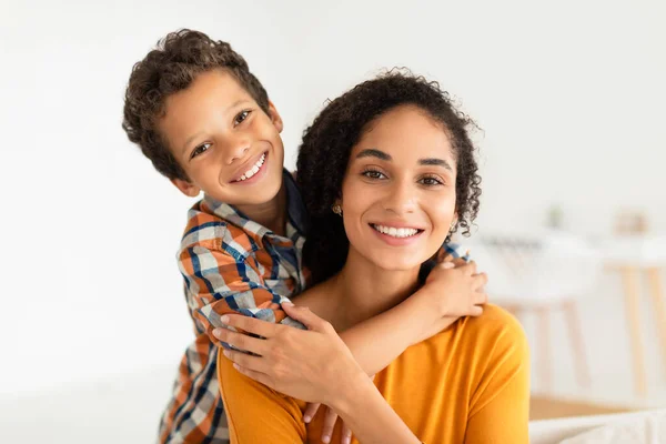 Portrait Of Latin Mom And Kid Son Sharing Warm Embrace Smiling To Camera At Home. Happy Boy Hugging His Mommy From Back Posing Together. Family Bonding, Joy Of Motherhood Concept