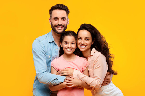Happy european parents hugging their pretty daughter and smiling to camera on yellow background, studio shot. Spouses embracing and posing with child girl