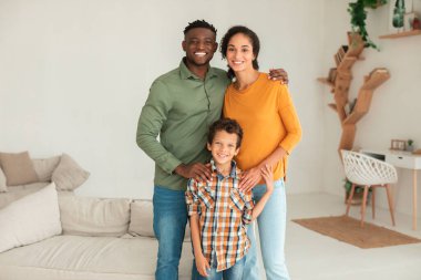 Farklı aileleri sevmek. Neşeli Afrikalı Amerikalı adam Arap karısını ve küçük oğlunu kucaklıyor, fotoğraf için birlikte poz veriyor, modern oturma odasında dururken kameraya bakıyor..
