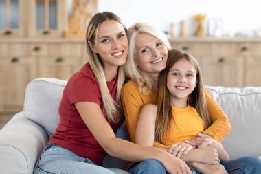 Mutlu, güzel, sarışın kadınların aile portresi. Üç kuşak anne, büyükanne ve çocuk evde. Güzel bir genç kadın, anne ve kızına sarılıyor, aile fotoğrafı için kanepede poz veriyor.