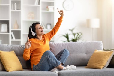 Neşeli, uzun saçlı, Hintli bir kadın oturma odasında oturmuş, yeni kulaklıklarının keyfini çıkarıyor, şarkılar söylüyor, kapalı gözlerle müzik dinliyor ve dans ediyor.