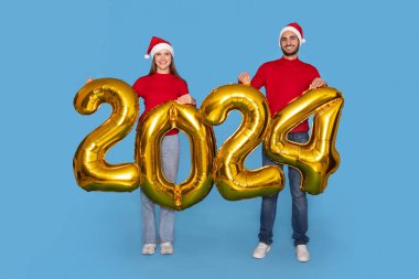 Noel Baba Şapkalı mutlu çift, ellerinde altın 2024 balon, kameraya gülümsüyor, Noel ve yeni yıl tatillerinde neşeli erkek ve kadınların uzun pozları, mavi bir geçmişi var.