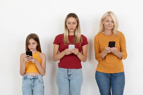 Gadgets addiction, lack of communication in family. Three generations women using phones, posing on white background. Adults mom and grandma and child teen daughter with modern gadgets smartphones