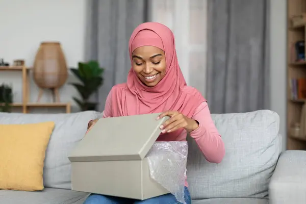 Happy african american muslim woman opening delivery box and looking inside, unboxing parcel with purchases while sitting on couch in living room interior, copy space