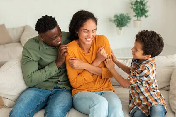Family Fun. Diverse Parents And Kid Boy Laughing And Cuddling While Dad And Son Tickling Mom, Sitting On Sofa Indoor. Man, Woman And Their Child Bonding Enjoying Weekend Together