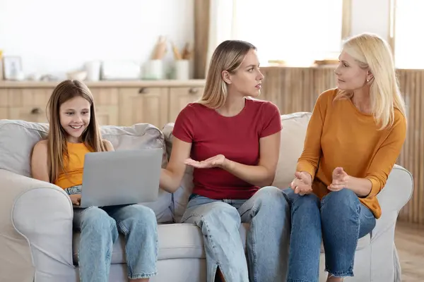 Gadget addiction for teenagers concept. Worried young woman mother complaining grandmother about teen daughter using laptop computer. Three generations family sitting on couch at home