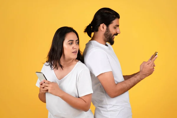 Shocked millennial arab woman looking at husband phone, isolated on yellow studio background. Online cheating, chat, family and relationship problems, mistrust, gadget addiction
