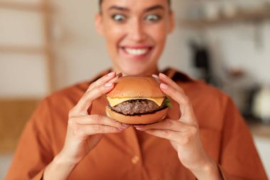 Fast food bağımlılığı. Burger tutan aç bir kadının portresi ve açık gözlerle ona bakmak, lezzetli atıştırmalıkların tadını çıkarmak, mutfakta oturmak, seçici bir odaklanma.