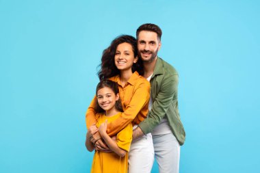 Üç kişilik mutlu bir beyaz aile. Kızı kameraya sarılıp gülümsüyor ve mavi arka planda poz veriyor. Avrupalı ebeveynleri ve kız çocuklarını seven