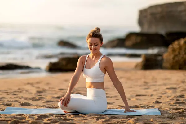 Young european lady athlete in sportswear sitting in lotus position practicing yoga, enjoying stretching body exercises on sea beach. Fit outdoor, sport and weight loss