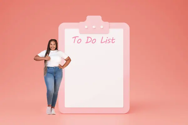 Cheerful young black lady in casual show thumb up near big plan with free space, do it list, isolated on pink studio background. Approval, recommendation future planning, motivation and weight loss