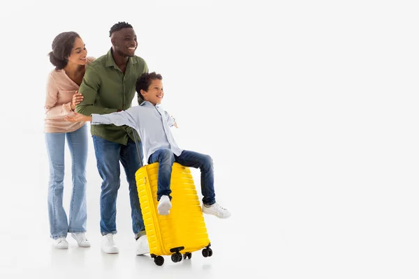 Family Vacation Trip. Joyful Diverse Parents Riding Their Son On Yellow Travel Suitcase On White Background. Boy Spreading Arms Like Plane Wings, Advertising Flight Tickets Sales Offer, Copy Space